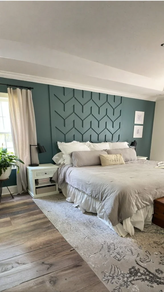 A deep teal accent wall featuring a custom geometric chevron-style molding design, illustrating elegant bedroom wall ideas. In this image, the textured wood paneling serves as a striking backdrop for a bed with ruffled white and grey linens, flanked by a white nightstand with a black modern lamp and a distressed light-patterned rug over hardwood floors.