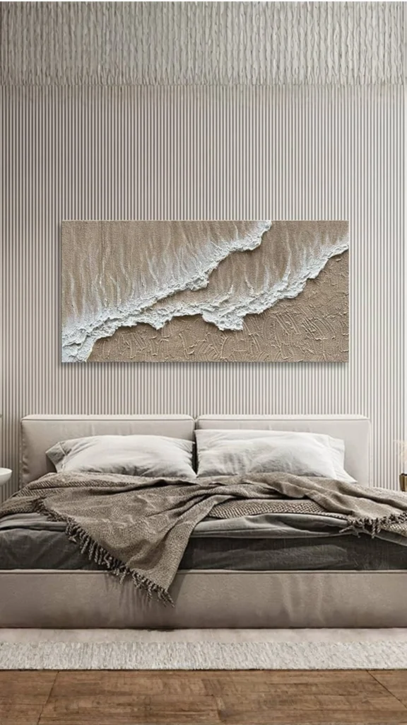 A large rectangular canvas with heavily textured plaster art depicting 3D white waves on a beige background, highlighting unique bedroom wall ideas. In this image, the artwork is mounted on a wall with fine vertical stripes above a modern beige bed styled with layered grey and brown textured blankets, creating a sophisticated and calming gallery-like feel.