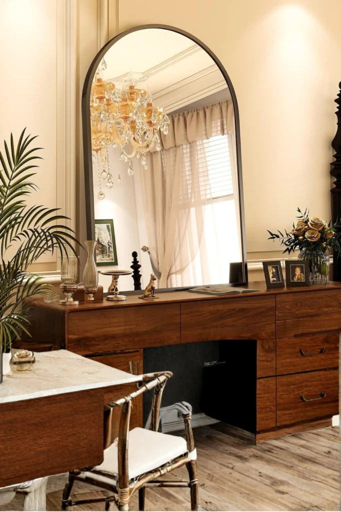 A luxurious bedroom vanity setup featuring a large arched tabletop mirror resting on a rich walnut wood dresser. The mirror reflects a crystal chandelier, beige curtains, and warm lighting. Small decorative frames, a gold statue, and glass vases style the top of the wooden desk, with a matching chair in the foreground. This elegant look shows how, to create a cohesive, high-end studio feel, focus on dedicated wood bedroom accessories. Add a small vanity mirror framed in warm oak or walnut to bring a soft, curved texture to your desk.