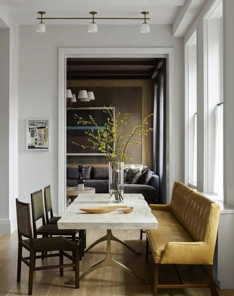 A luxury dining nook maximizing an open floor plan with a built-in upholstered banquet bench in a warm mustard yellow tone, placed neatly against a crisp white wall under a large window. A rectangular white marble dining table sits on light wood flooring, paired with three dark wood chairs with olive green seats. The inviting layout flows naturally into an adjoining living room in the background, which features a moody dark gray sofa and a large abstract art piece.