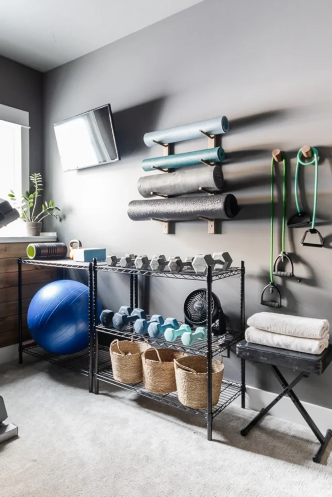 A beautifully organized home workout space featuring creative room wall ideas that maximize floor space. Mounted on a matte grey wall are a wooden rack holding layered yoga mats, a TV, and wall pegs with resistance bands. Below, a black wire shelving unit neatly holds dumbbells, a blue stability ball, a small fan, and woven storage baskets, perfectly showing how to introduce functional design elements that double as home decor.