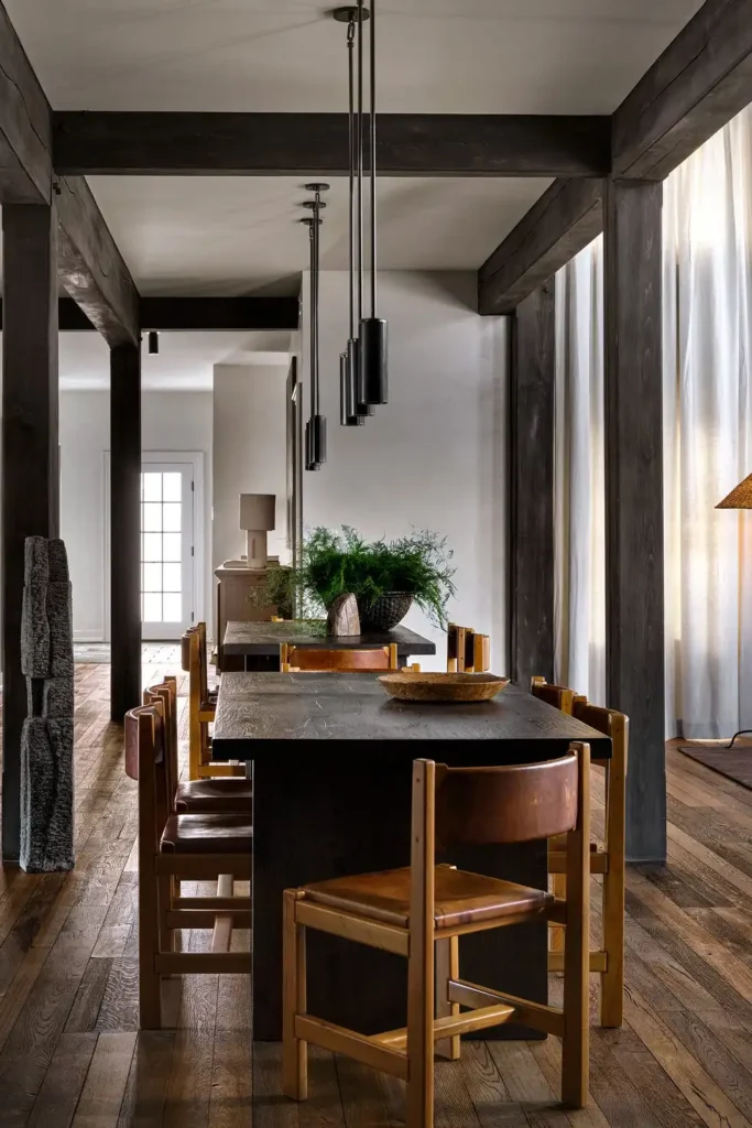 A rustic, high-end dining area displaying an authentic designer look where the architectural layout blends seamlessly with your furniture. The space features long, heavy dark wood dining tables paired with mid-century modern wooden chairs with tan leather backrests under sleek, vertical black cylindrical pendant lights. Heavy, dark timber support columns and ceiling beams frame the room, while a soft neutral beige wall stands in the background, creating a balanced backdrop for elevated wall dining room decor.