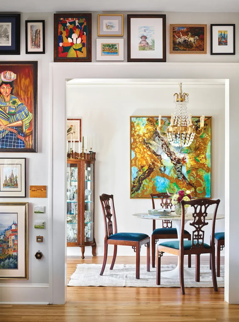 A vibrant, maximalist dining room viewed through a large white doorway surrounded by an eclectic salon-style gallery wall of framed paintings, showcasing creative gallery wall ideas. In this image, the adjacent room features a marble-top dining table with high-backed wooden chairs, a sparkling crystal chandelier, and a large, colorful abstract painting in shades of gold and turquoise mounted on the main wall.