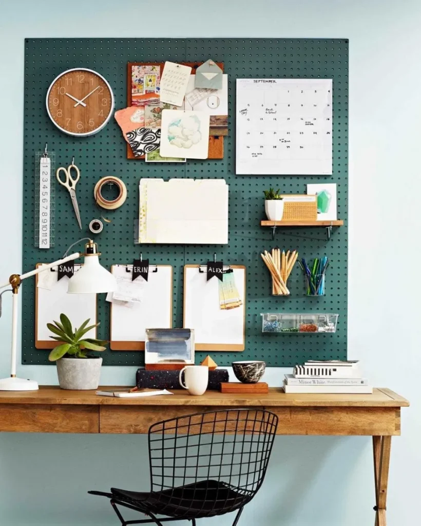 A functional and organized home office setup featuring a large dark green pegboard mounted on a pale blue wall, demonstrating practical office design ideas. In this image, the grid organizer holds an analog clock, a calendar, clipboards, scissors, and small shelves with supplies and a tiny plant, sitting above a warm wooden desk with a wire back chair and a white task lamp.