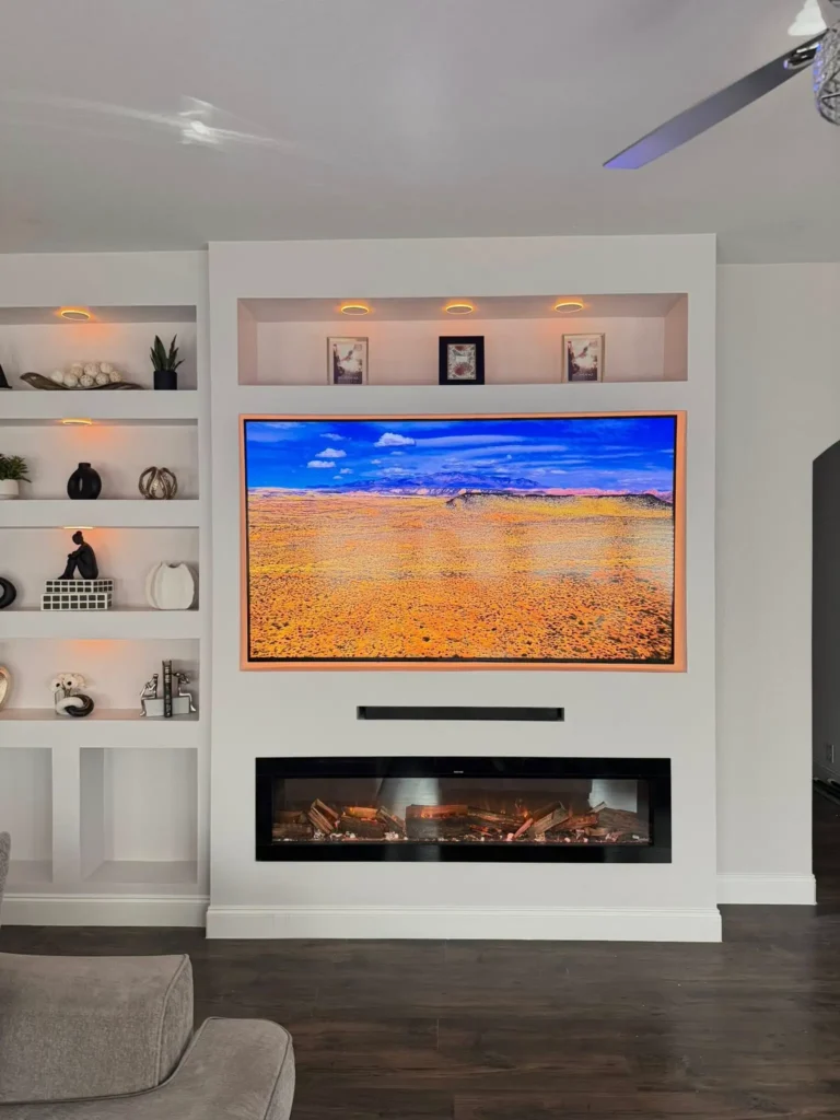 A modern white custom built-in entertainment center featuring a recessed flat-screen television with warm backlit lighting, situated above a linear electric fireplace and flanked by illuminated display shelving.