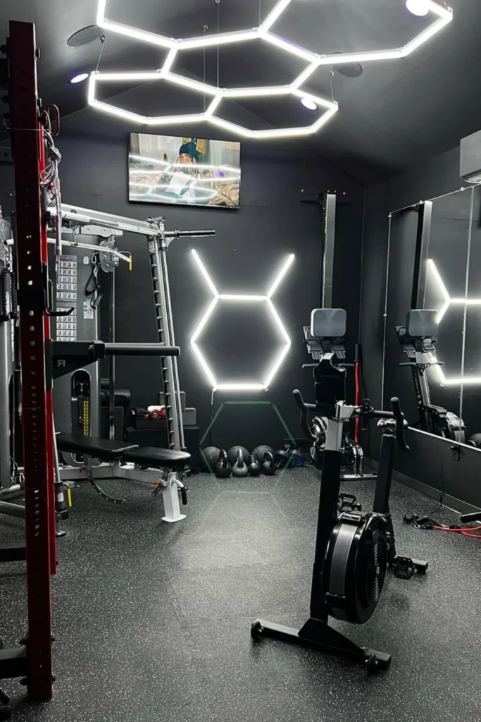 A futuristic home gym with a matte black theme, showcasing bright white geometric hexagonal LED light fixtures on the ceiling and a matching neon wall light fixture glowing in the center of the room. The space is equipped with professional training gear, including a rowing machine, spin bike, smith machine cage, and a wall-length mirror reflecting the ambient lighting setup.