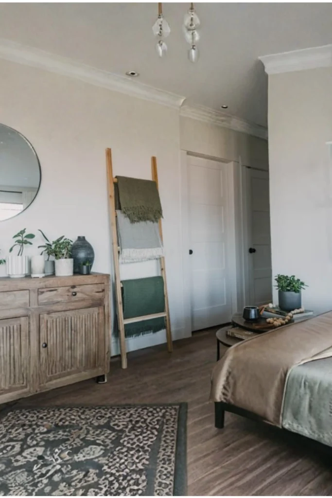 A cozy bedroom layout featuring a rustic wooden leaner blanket ladder resting against a plain white wall. The ladder holds three neatly folded throw blankets in earth tones: olive green, grey, and forest green. To the left, a wooden credenza is styled with potted plants and a round wall mirror, while a bed with neutral linens sits to the right on dark wood-look flooring with a patterned rug. This scene shows why diving into diy wood accessories home decor projects is a fantastic, budget-friendly way to style your space, especially when you build or style a simple wooden ladder against an empty wall to hang your favorite cozy throw blankets.