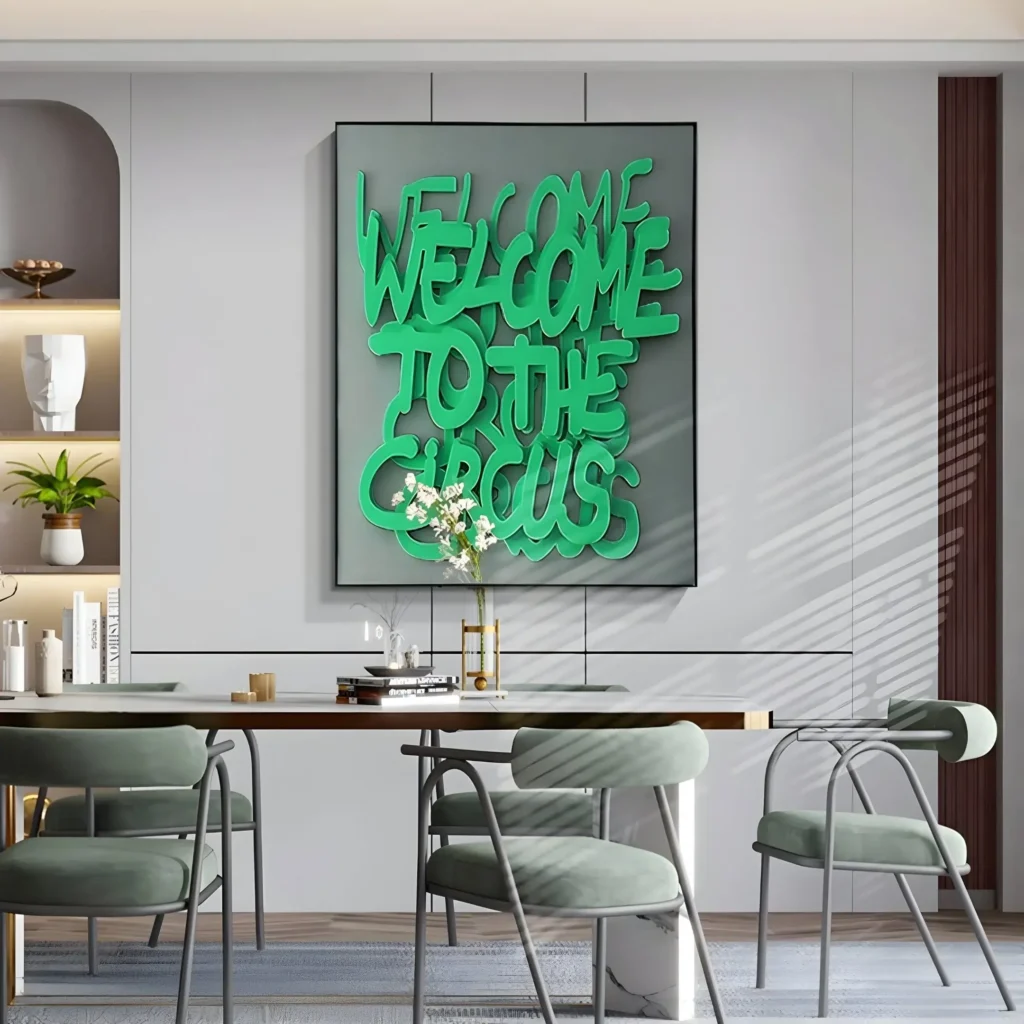 A modern dining area featuring a massive typography art piece in a large black frame mounted on a minimalist grey paneled wall. The single oversized frame showcases layered, bold green 3D lettering spelling out "Welcome to the Circus" on a solid grey background. Below the artwork, a contemporary dining table is surrounded by sleek, light green upholstered chairs, styled with books and a small white floral arrangement, under soft window blind shadows.