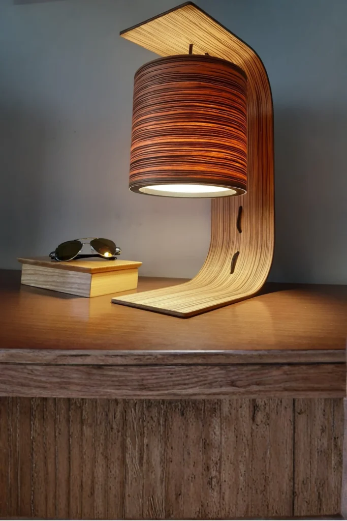 A modern wooden desk lamp with a curved C-shaped body and a cylindrical wood-grain shade casting a warm glow onto a wooden tabletop. In the background, a pair of sunglasses rests on a small light-colored wooden box against a neutral wall. This sleek design shows why you should choose nightstand lamps with solid wooden block or cylinder bases to add instant architectural interest to your bedside layout, keeping finishes matte to let the natural grain shine through.