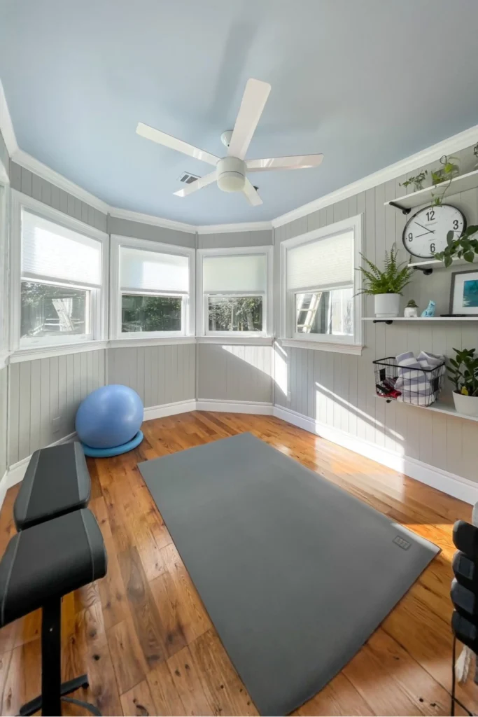 A light-filled home fitness space illustrating a gym room at home small spaces minimalist setup. A premium gray exercise mat sits centered on rich hardwood flooring under a soft blue ceiling with a white fan. The room features gray shiplap walls, a bay window, a blue stability ball, black workout benches, and floating wall shelves styled with plants and a large clock.