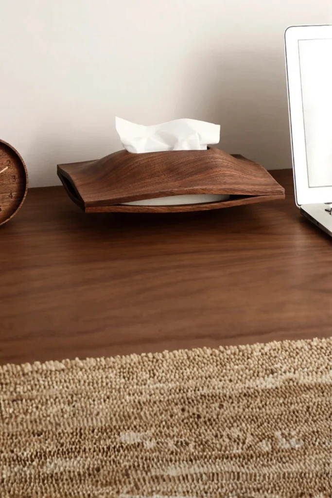 A uniquely shaped, curved wooden tissue dispenser holding white tissues on a rich brown wooden tabletop. The modern, minimalist piece has an organic wave-like design with a matte finish that highlights the natural grain. A small wooden desk clock is partially visible on the left, a silver laptop rests on the right, and a textured woven rug sits in the foreground. This layout elegantly shows how you can swap out bright, clinical store packaging for a minimalist matte wood tissue box cover that blends seamlessly with your nightstand furniture.