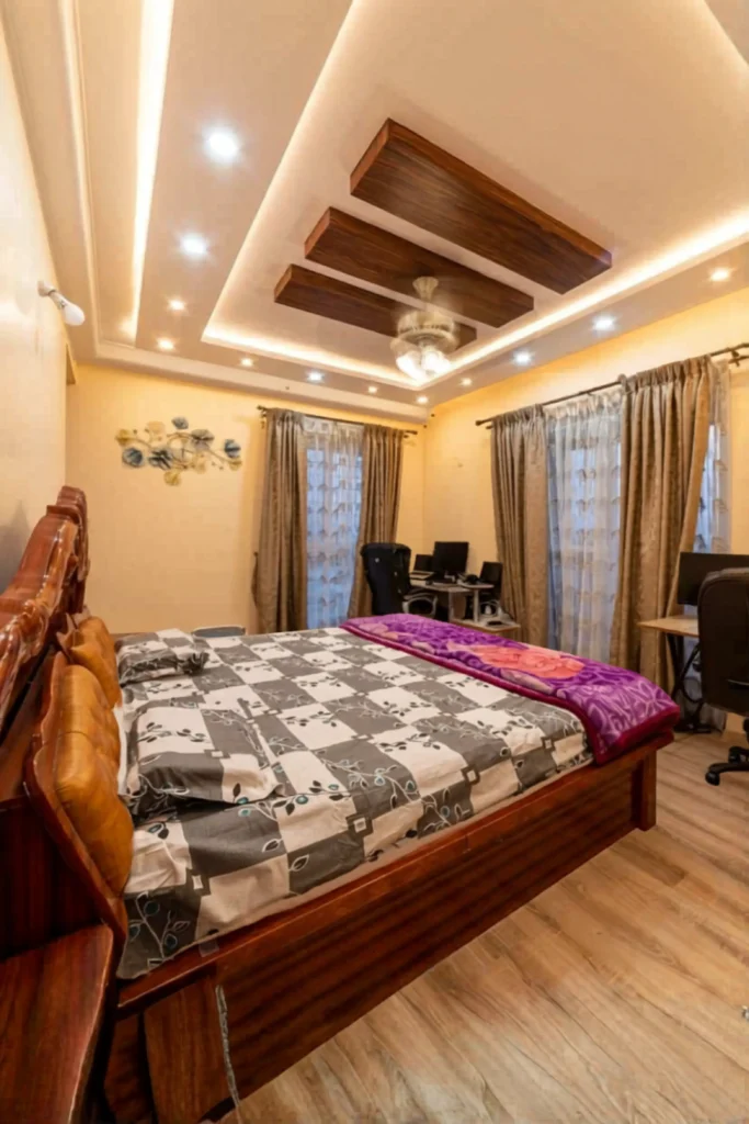 A bedroom layout highlighting creative bedroom ceiling ideas modern interior design simple inspiration. The tray ceiling features multi-tiered recessed paneling accented with warm built-in spot lighting and a decorative center wood panel design holding a glass ceiling light. Below, a large wooden bed with patterned sheets and a purple throw blanket rests on wood floors, framed by warm beige walls, long curtains, and small home office desk setups in the background.