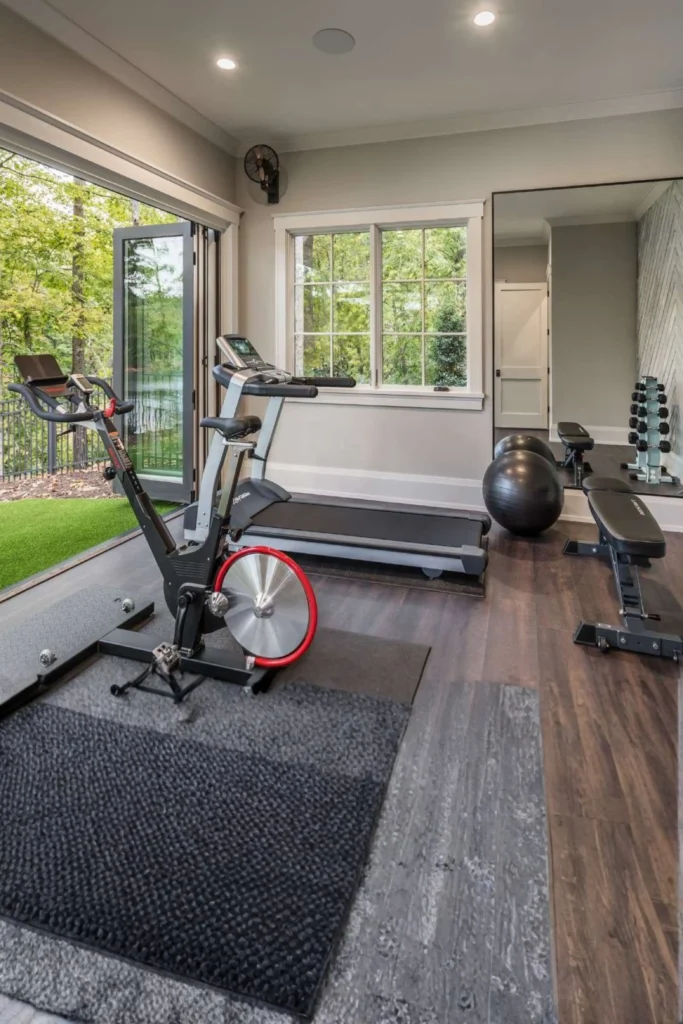 A light-filled home gym layout highlighting a seamless connection to nature, perfect for a gym room at home small spaces outdoor routine. A stationary spin bike with a red wheel accent sits on a textured dark gray rug, flanked by a treadmill under a window and a large wall mirror reflecting workout benches. Large glass folding doors open completely to a lush, green outdoor zone featuring vibrant artificial turf and surrounding trees.