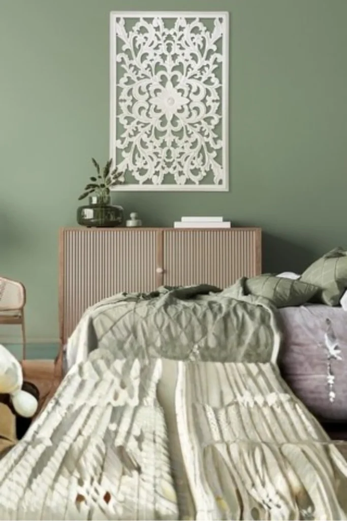 A large, rectangular white intricately carved wooden wall medallion hanging on a sage green accent wall. Below the artwork, a fluted light wood credenza stands behind a bed layered with textured sage green and neutral blankets and pillows. A dark glass vase with dried botanicals sits on top of the cabinet under soft lighting. This layout shows why you don't be afraid to use a single, massive wooden carved medallion above your headboard to act as a dramatic, jaw-dropping focal point on an empty accent wall.