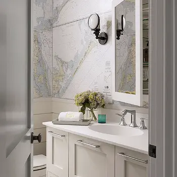 A classic, bright guest bathroom featuring light neutral wallpaper designed with an intricate, vintage nautical map pattern, showcasing clever guest bathroom wall decor ideas modern travelers will love. In this image, the sophisticated map accent wall serves as a backdrop for a white vanity with silver fixtures, a recessed medicine cabinet mirror, a black industrial wall sconce, and a small vase of hydrangeas.