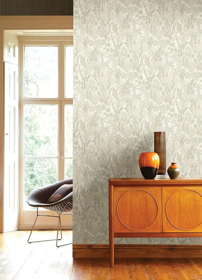 A bright room featuring a wall covered in a light beige and cream botanical peel-and-stick wallpaper with a dense leaf pattern, highlighting versatile wallpaper ideas. In this image, the patterned wall serves as a backdrop for a mid-century modern wooden credenza styled with decorative ceramic vases, next to an open doorway with soft natural light streaming onto the hardwood floors.