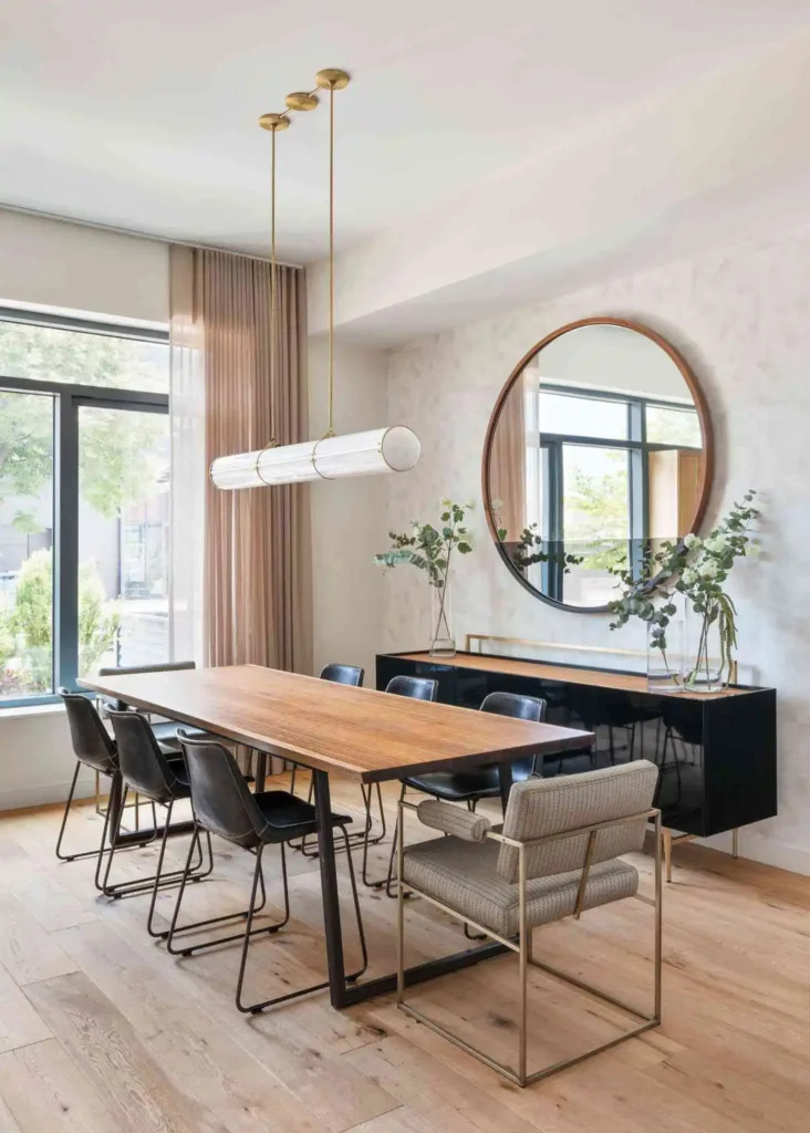 A bright, upscale dining room featuring a striking wall dining room mirror arrangement with a large, round wooden-framed mirror centered above a glossy black credenza sideboard. The mirror features a dual-tone or half-tinted glass panel that elegantly reflects the large window across from it, maximizing natural light. In the foreground, a long natural wood dining table is surrounded by sleek dark leather chairs and a textured armchair, positioned under a modern linear cylindrical pendant light.