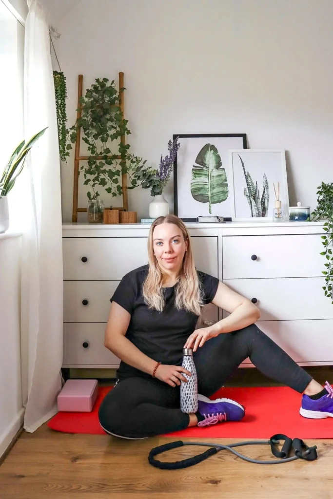 A woman sitting on a bright red yoga mat holding a water bottle, showcasing a dedicated self-care and recovery station designed to restore your body and mind. She is leaning against a large white dresser styled like a luxury spa setup with framed botanical art, a reed diffuser, fresh lavender, and potted hanging plants on a wooden ladder. A pink yoga block and a black resistance band rest on the wood floor nearby.