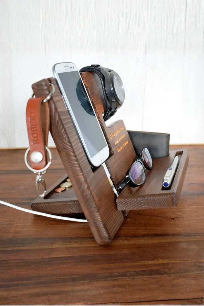A dark brown wooden docking station and valet stand resting on a matching wood tabletop. The stand holds a smartphone with a white charging cable connected, a black wristwatch, a pair of eyeglasses, a pen, a wallet, and a leather keychain engraved with "ROBERT". This functional layout beautifully showcases how to use wood accessories home decor modern elements to keep daily essentials organized in a fresh and open space.