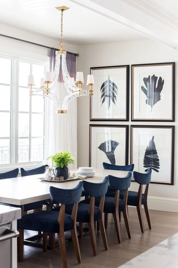 A bright, elegant dining room showcasing a structured, balanced grid gallery wall with four identical dark-framed prints. The chosen dining area wall art features minimalist black-and-white botanical sketches with wide white paper mats, hung with equal spacing to create a dynamic, expensive look. In the foreground, a light wood dining table is surrounded by deep blue upholstered chairs beneath a modern glass and gold chandelier hanging near a large window.