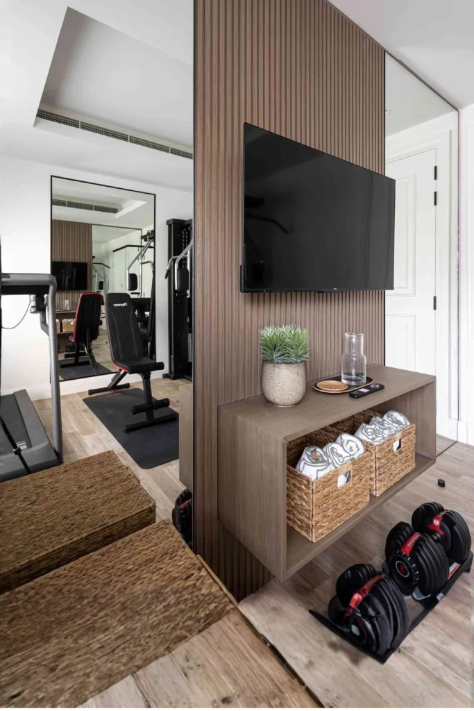 A multi-purpose room setup showing how to use multi-functional furniture to hide your gym equipment. A slatted wood room divider panel with a mounted TV features a floating wooden console shelf beneath it holding a potted plant, water carafe, and woven baskets filled with towels. On the light wood floor, a pair of adjustable dumbbells rests neatly out of the way, while large floor-to-ceiling mirrors reflect a hidden workout space with a treadmill and adjustable bench behind the partition.
