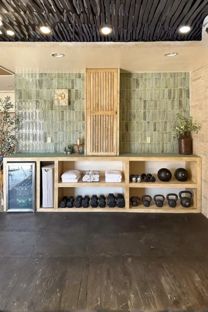 A stylish home fitness zone showcasing smart at home gym room small spaces ideas that maximize vertical real estate. Against a green textured tile accent wall, a custom wooden built-in shelving unit keeps the dark workout floor completely clear for movement, neatly organizing dumbbells, kettlebells, medicine balls, and white towels. A tall wooden slatted cabinet rests on the counter alongside a small beverage fridge.