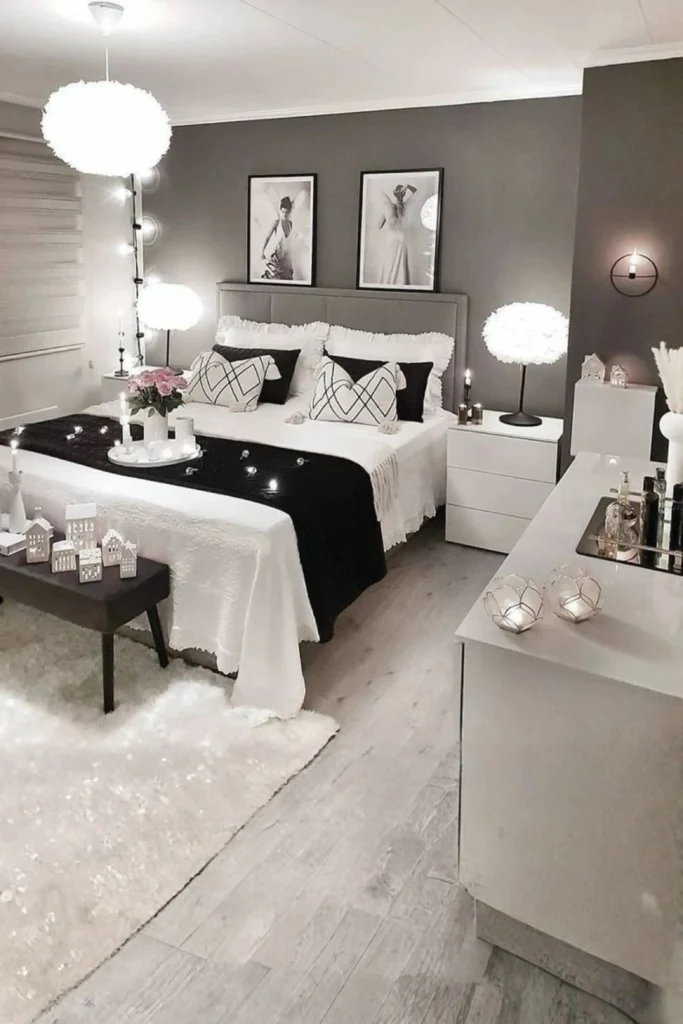 Ambient Mood Lighting layers in a modern gray and white bedroom setup. The image shows a beautifully styled bed with geometric accent pillows and a black bed runner adorned with small fairy lights. Fluffy, white cloud-like lamps sit on white nightstands as task lighting on both sides of the tufted gray headboard, complemented by a matching statement pendant ceiling light and warm candle accents on a white dresser in the foreground.