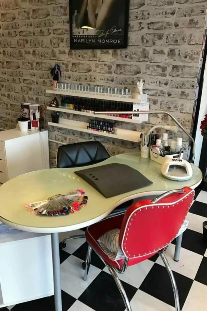 A beautifully organized manicure setup showcasing professional beauty room ideas salon at home nails stations. The image features clear acrylic shelves mounted on a light beige wall, neatly displaying rows of vibrant gel nail polish bottles organized by shade. On the white desk below sits an adjustable gold task lamp, creating a clean and functional home salon workspace.