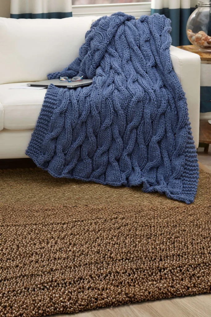 An up-close shot of a white sofa beautifully draped with a heavy, oversized knit blanket in a dusty blue color, showing off its thick chunky cable-knit pattern. The large blanket cascades down the couch cushions onto a highly textured, brown woven jute area rug below, giving the living room layout an immediate visual warmth.