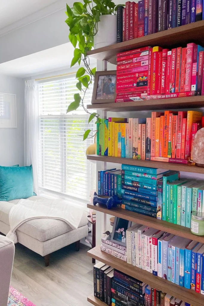 A vibrant home bookcase showcasing stunning color-coded rainbow shelves to style books by color. The open wooden shelving unit displays rows of books organized beautifully into a seamless color gradient, transitioning smoothly from reds and pinks down to yellows, greens, and deep blues. A green trailing pothos plant sits on the top shelf, while a cozy cream chaise lounge chair rests next to a bright window in the background.