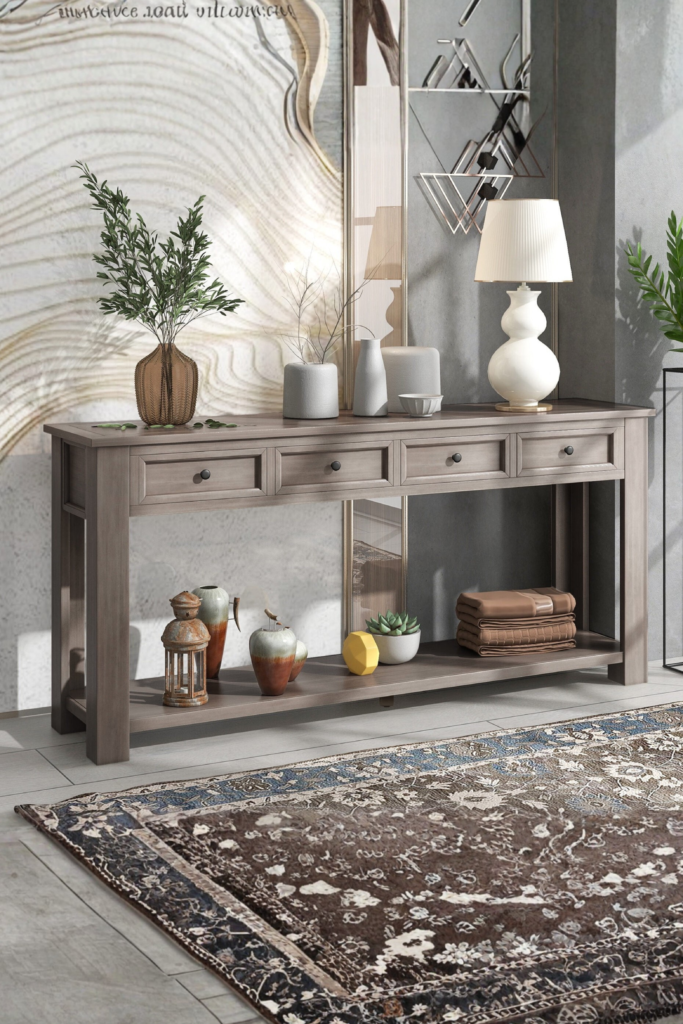 A long, rustic gray wooden console table with drawers and a bottom storage shelf positioned in an elegant entryway. The tabletop is decorated with a white structural table lamp, minimalist ceramic vases, and greenery, while the bottom shelf holds folded towels and decorative pottery over a vintage patterned area rug, perfectly illustrating timeless furniture ideas for new home styling.