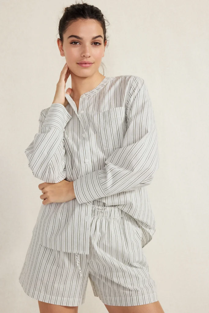 A young woman with her hair tied back in a bun posing against a neutral cream-colored wall. She is wearing a matching striped cotton poplin lounge set featuring an oversized, collarless long-sleeved button-down shirt with a breast pocket and matching boxer-style shorts with an elastic drawstring waist. This effortless, clean-girl aesthetic is a timeless look that is perfectly structured and breathable for staying comfortable at home.