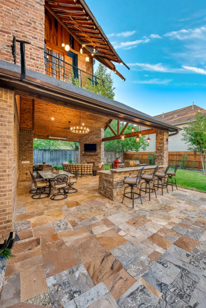 A spacious luxury covered patio with a wooden ceiling and stone flooring, featuring an outdoor dining set, a backyard kitchen island with bar stools, and a cozy seating area under a glowing chandelier.