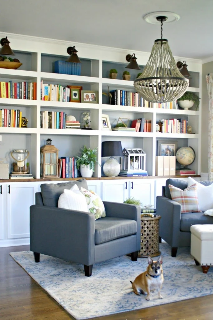 A stunning living room featuring home book library ideas styled with a large white built-in bookcase. The shelves are artfully arranged with a mix of colorful books and curated decor items like small green plants, decorative lanterns, and a vintage globe to tell the story of who you are. In the foreground, two comfortable grey armchairs rest on a patterned blue area rug under a unique beaded chandelier lamp, with a small dog sitting near the center.