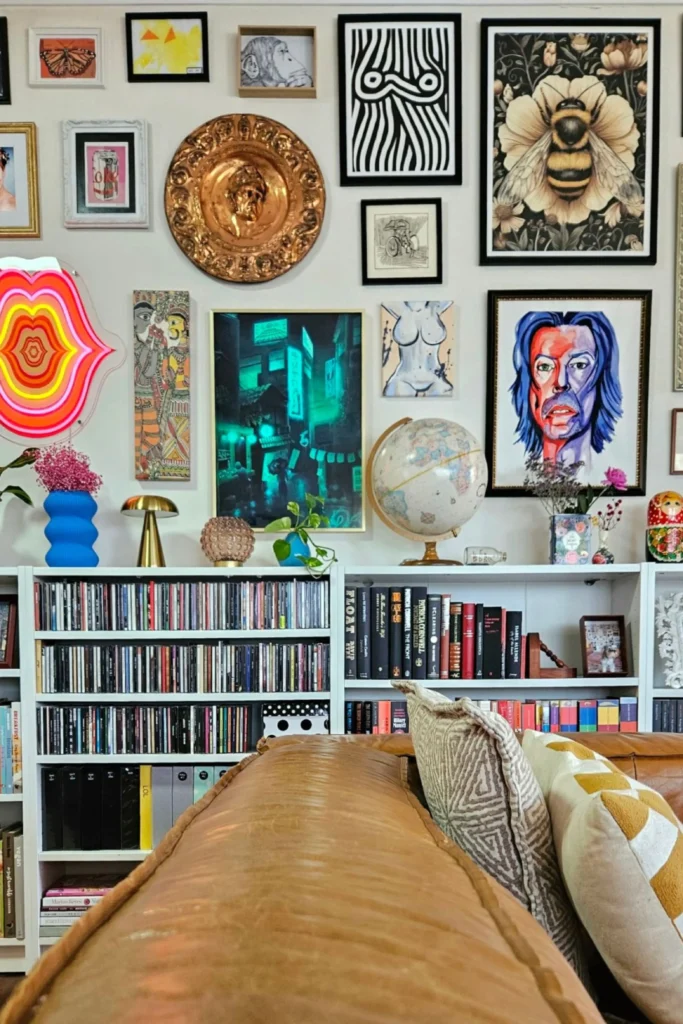 An intentionally curated maximalist display featuring white shelves packed with books and a media collection behind a tan leather sofa. The white wall above the shelves serves as a dense gallery wall showcasing an eclectic mix of art prints, a large metallic brass plate, an abstract neon lips light, and a vintage desk globe.