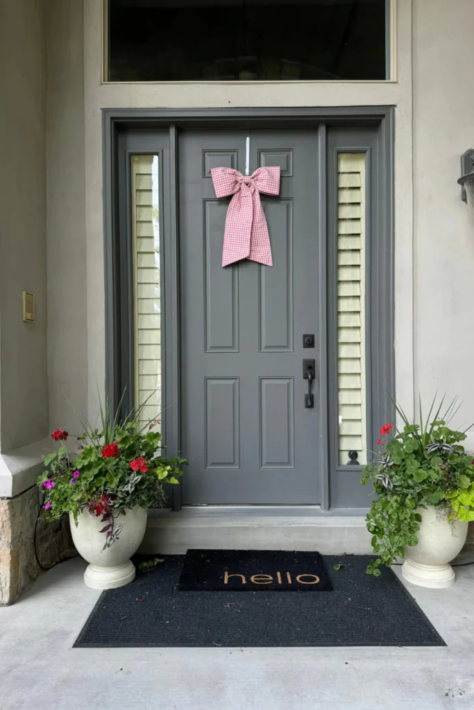 A clean and beautifully organized front porch entryway featuring a sleek charcoal gray strong front door as a fresh canvas. The paneled gray door is decorated with a pink gingham bow and flanked by narrow sidelight windows with cream shutters. Two large white urn planters filled with vibrant green plants and red flowers stand on a swept concrete porch on either side of a black "hello" doormat, showcasing welcoming gray strong front door ideas.
