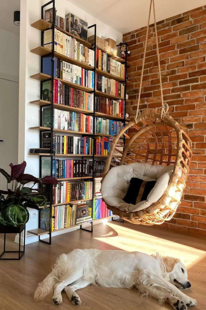 A cozy reading corner highlighting comfortable home library ideas book lovers adore. The space features a unique rattan hanging egg chair with plush tufted cushions suspended next to an exposed red brick accent wall. A tall, modern black metal bookshelf tightly packed with colorful books stands in the background, illuminated by natural sunlight splashing across the wooden floor where a golden retriever dog is peacefully napping.