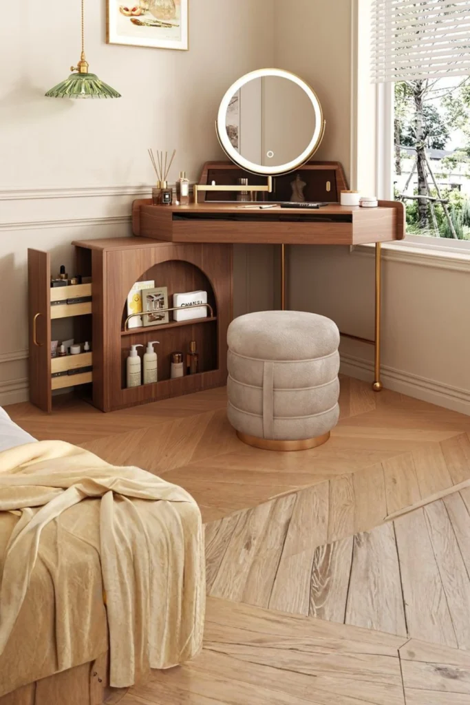 A luxury small-space dedicated vanity nook set on a raised wooden floor next to a bed. The setup features a sleek, compact walnut wood vanity desk with gold metal legs, an illuminated round LED mirror, and a matching beige tufted storage stool. The desk includes a unique pull-out side organizer cabinet displaying skincare, makeup products, and books, maximizing hidden storage perfectly.