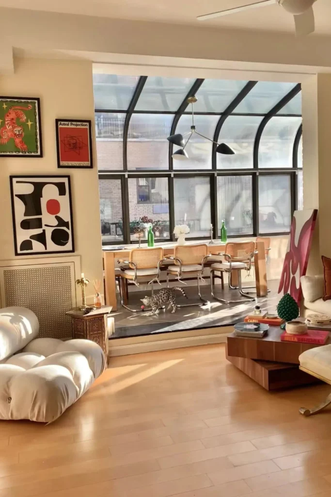 A sunlit apartment interior highlighting smart studio apartment layout design ideas home office setups. The space features a chic living zone with a plush white bubble chair and wooden coffee table, transitioning into a dining and workspace area in the background with cane chairs set against a dramatic glass atrium window.