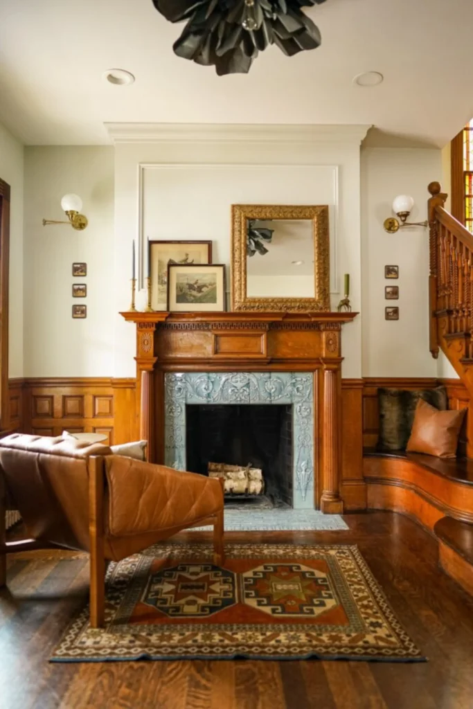 A warm living space demonstrating how breaking classic Victorian interiors symmetry creates a contemporary edge, inspired by Ditch Symmetry for a Modern Vibe.jpg. Features include a grand wooden fireplace mantel with intricate light blue tiling, an ornate gold mirror, a single brown tufted leather armchair on an patterned area rug, and an asymmetrical built-in wooden bench seat tucked under a staircase.