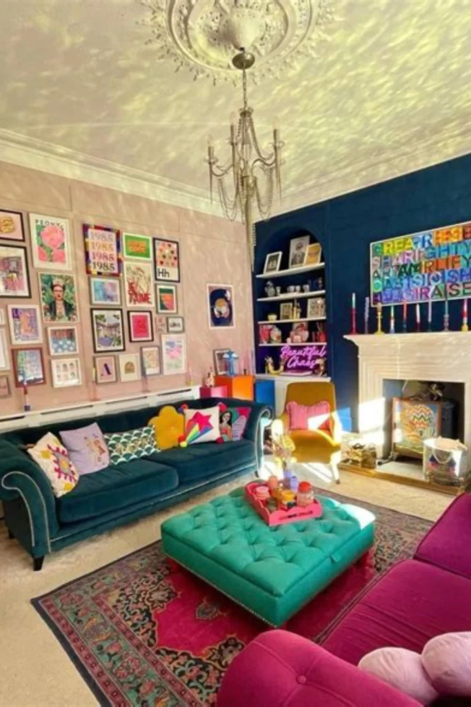 A vibrant maximalist living room showcase from the image "Dopamine Decor.jpg". The room features a deep teal tufted chesterfield sofa adorned with multicolored eclectic throw pillows, paired with a bright turquoise tufted ottoman sitting on a rich magenta patterned area rug. The walls are split into a soft blush pink gallery wall packed with colorful art prints on the left, and a dark navy blue accent wall on the right with a white fireplace mantle, a neon sign reading "Beautiful Chaos," and a mustard yellow armchair tucked into the corner under an ornate crystal chandelier.