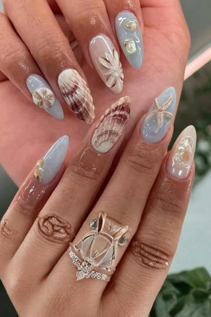 A close-up showcasing an intricate, beach-inspired 3D manicure featuring detailed designs of starfish, 3D seashells, and delicate pearls encapsulated onto glossy stiletto and almond-shaped nails. This artistic ocean theme highlights beautiful nail accessories design inspiration on how to style fragile, organic natural elements perfectly.