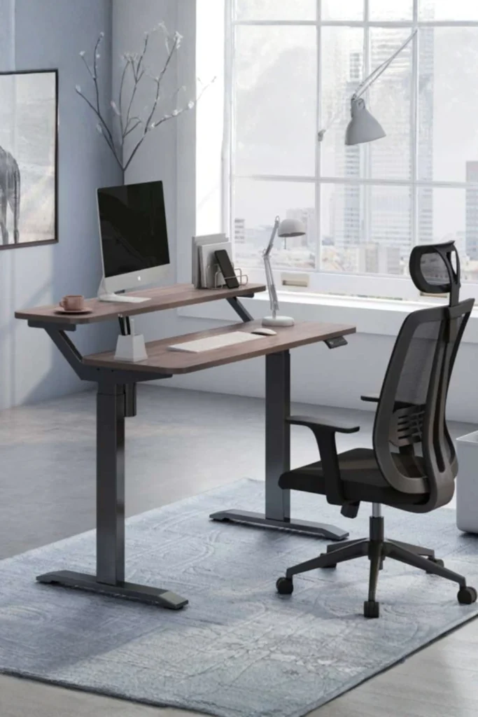 A modern home office featuring an ergonomic adjustable desk with a dual-tier wooden top, black metal frame, and a desktop computer monitor. Paired with a high-back black mesh office chair on a textured gray area rug next to a large window overlooking the city, demonstrating how creating a productive workspace is essential for modern living.