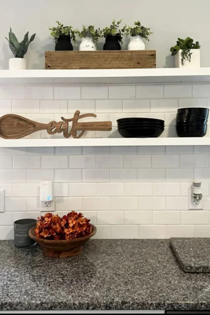 Kitchen counter area with white subway tile backsplash, white floating shelves displaying black bowls, a wooden "eat" sign spoon, and several small artificial plants in planters.