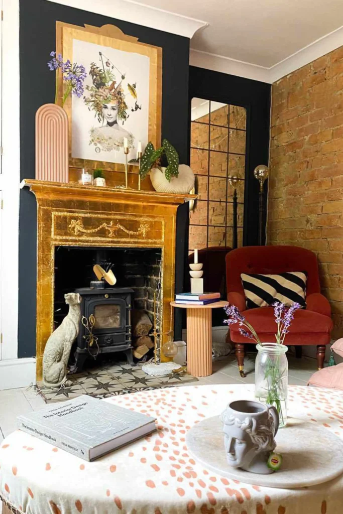 A layered eclectic living room featuring victorian decorating ideas diy projects inspired by Focus on Craft.jpg. The space showcases a distressed gold-leaf fireplace mantel with an inset black log stove cooker, a grey dog statue, and an exposed brick accent wall. A plush rust-red armchair with a striped pillow sits next to a fluted pink side table, with creative decor and art adding handmade texture.