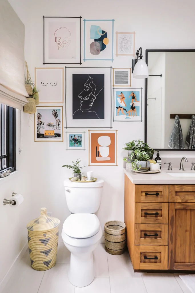 A bright modern bathroom gallery wall featuring an eclectic mix of abstract line art, colorful prints, and coastal illustrations taped directly to the wall above a white toilet, reminding you: Don't neglect your bathroom walls! Hanging a set of framed botanical art prints or vintage sketches brings a touch of nature indoors. The vanity area includes a wooden cabinet with drawers, a large mirror, potted green plants, and a white wall sconce. When paired with soft lighting, these art pieces transform a plain wall into a beautiful focal point that enhances the cozy, country-cottage aesthetic, complete with a woven laundry basket on the floor.