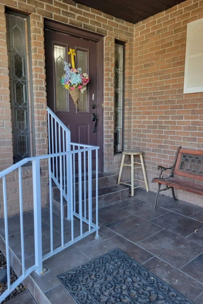 A brick front porch entryway highlighting common front door challenges, featuring a dark mauve or plum colored door with inset decorative glass panels and a hanging floral basket arrangement. The door is flanked by two long sidelight windows with detailed leaded glass geometric patterns, showcasing how balancing visibility with safety is a major priority. A white metal safety handrail leads up the tiled steps, while a wooden bench and a small stool sit against the brick wall next to a patterned black rubber doormat.