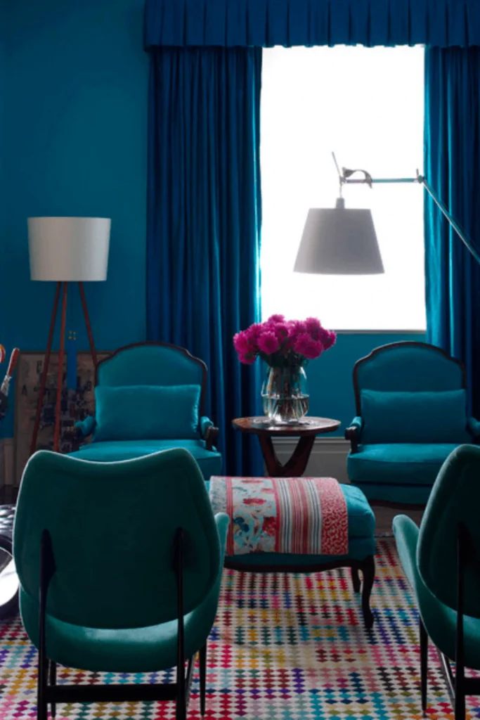A vibrant living room showcasing rich, dramatic, and moody color stories inspired by Go for Bold Colors.jpg. The space features deep sapphire blue walls with matching long curtains framing a bright window. Two teal tufted armchairs sit in the background, paired with modern teal chairs in the foreground, a colorful patterned rug, and a central small table with bright pink flowers.
