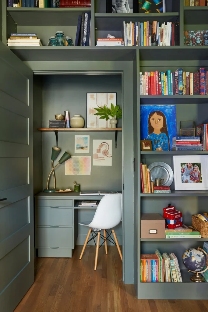 A clever deep green closet converted into a cozy built-in desk and reading nook, showcasing smart home book library ideas. Inside the opened closet doorway, a sleek matching green desk holds a modern white chair with wooden legs, an adjustable desk lamp, and framed watercolor artwork on the wall. The surrounding wall is lined with floor-to-ceiling built-in bookshelves densely packed with colorful books, kids' toys, and a vintage globe.
