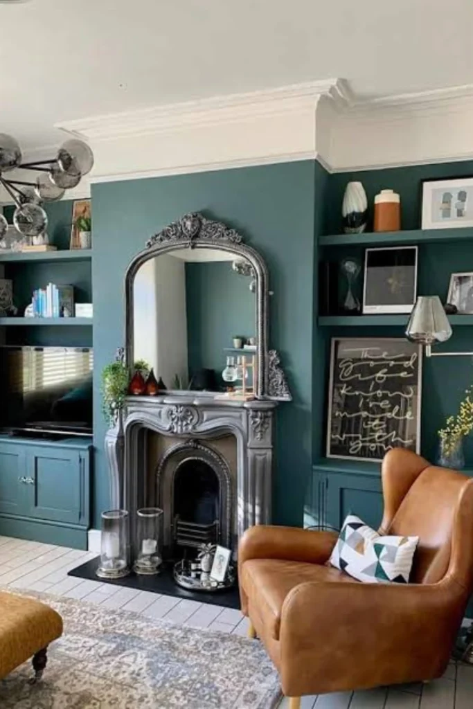 A personalized living room showing how a home can look like a reflection of you, not a replica, inspired by Include Your Own Personal Style.jpg. Features include a classic grey ornate fireplace mantel with a matching arched mirror, deep teal green walls with built-in bookshelves, a comfortable tan leather wingback armchair with a geometric pillow, and unique modern decor pieces that showcase a personalized, timeless aesthetic.